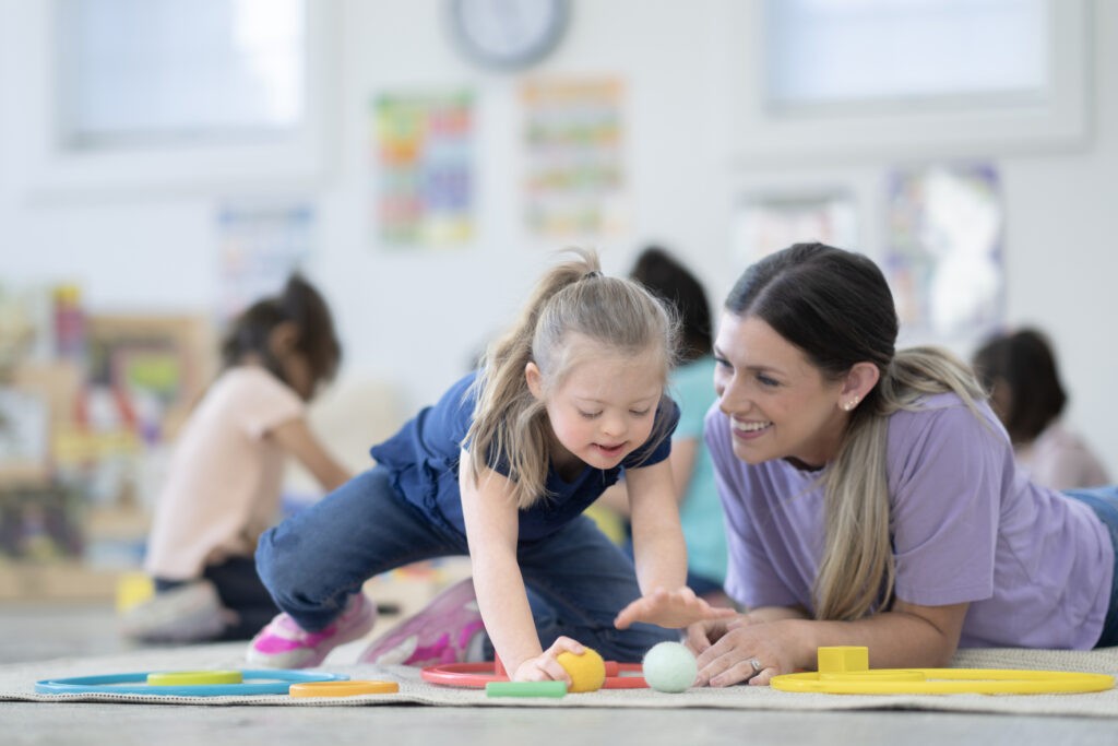 A sweet little girl with Downs Syndrome works with her teacher on the floor of the classroom as they short toys by colour. The two are both dressed comfortably and focused on the task as they work together.